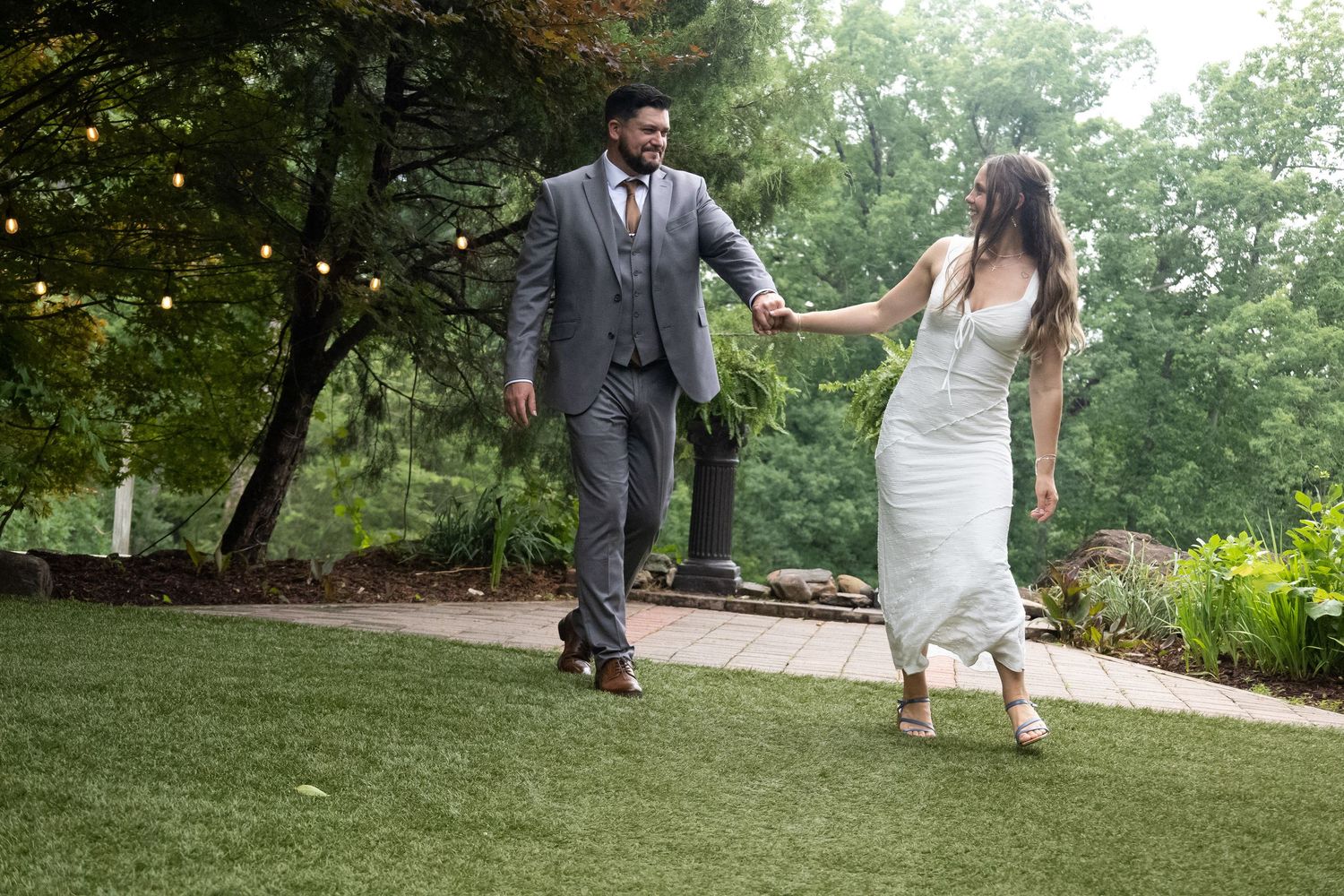 Bride and groom holding hands and dancing joyfully on green lawn surrounded by trees with string lights.