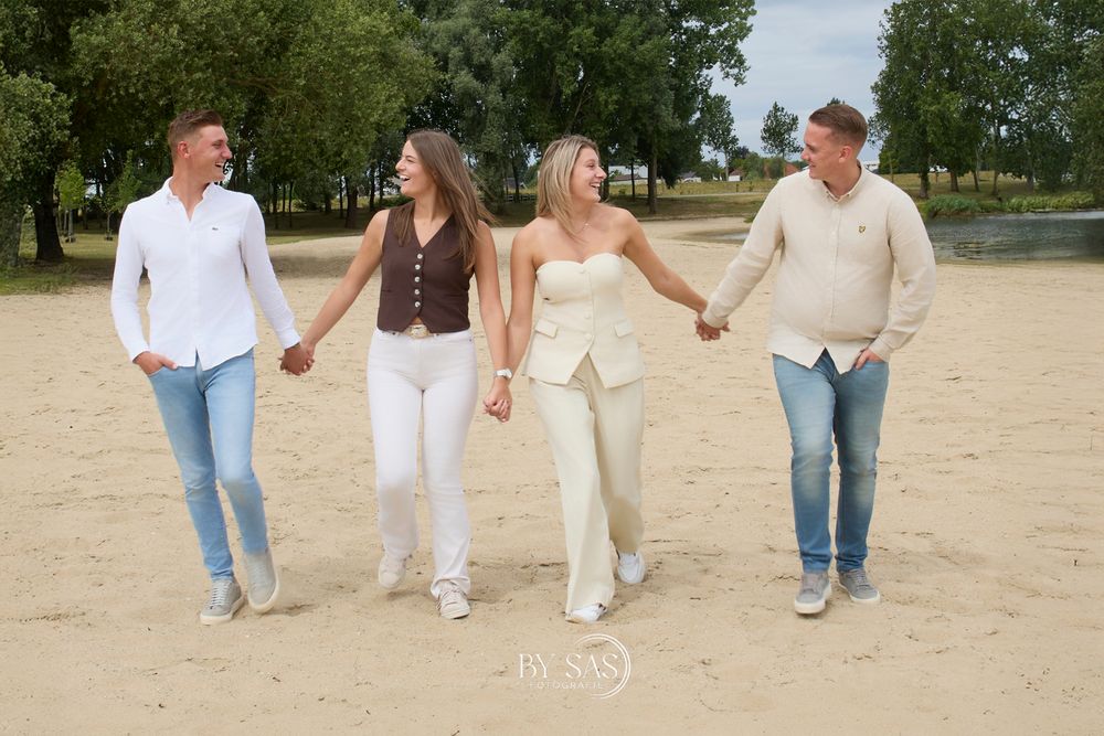 Four friends holding hands walk on sandy beach, two couples in casual neutral outfits smiling on a sunny day.