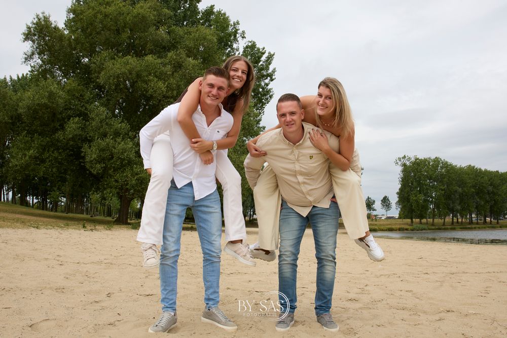 Two couples pose on a sandy beach, men giving women piggyback rides, with green trees in the background.
