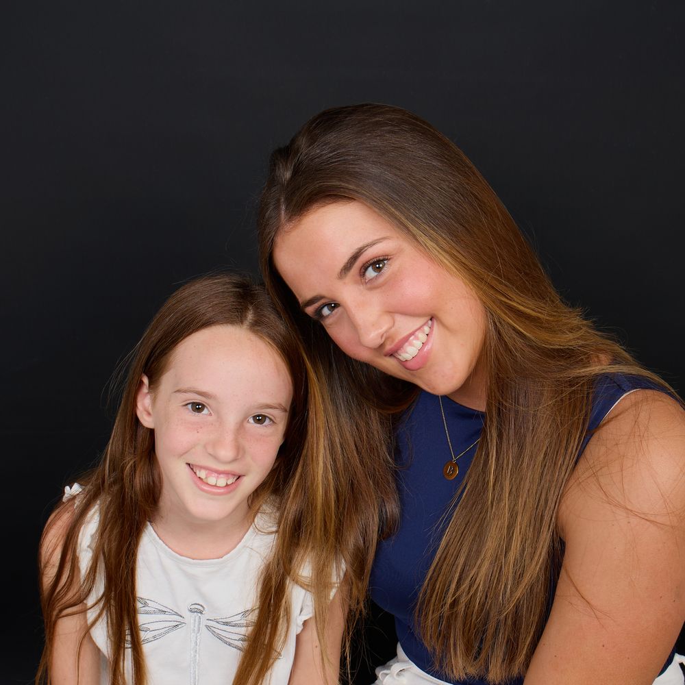 Two smiling girls with long brown hair pose together against a dark background in a portrait photo.