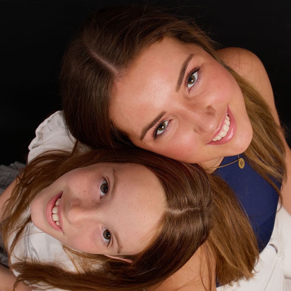Two smiling young girls with brown hair posing together against a dark background in a close portrait photo.