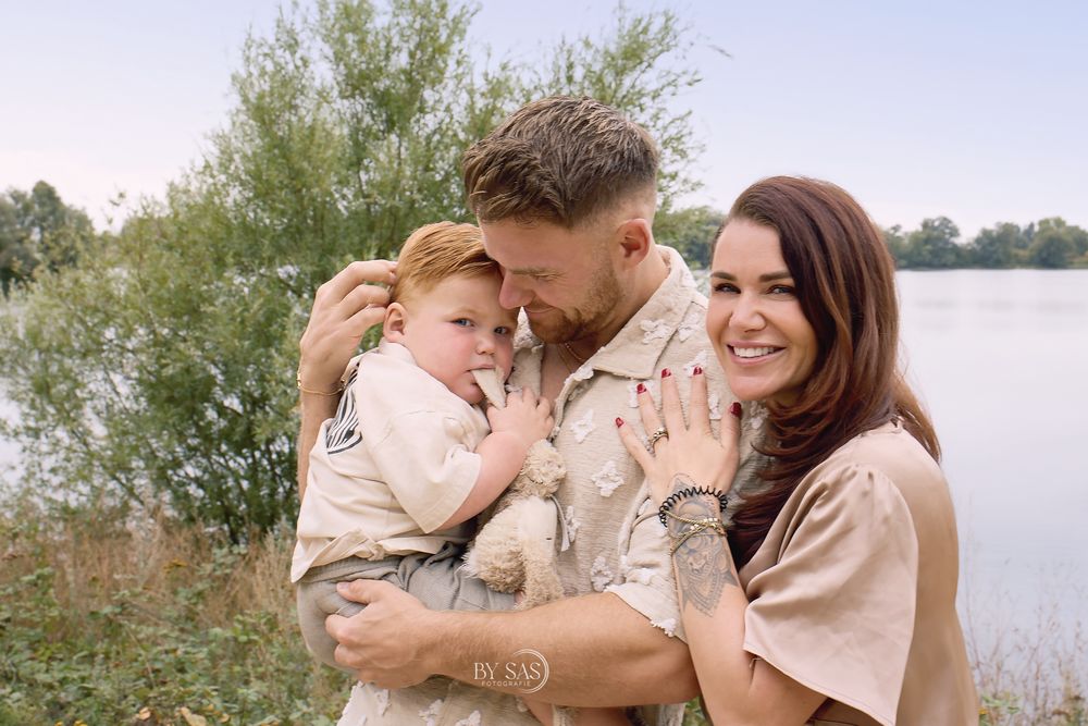 Military family portrait with soldier holding toddler beside smiling woman near a lake with lush greenery.