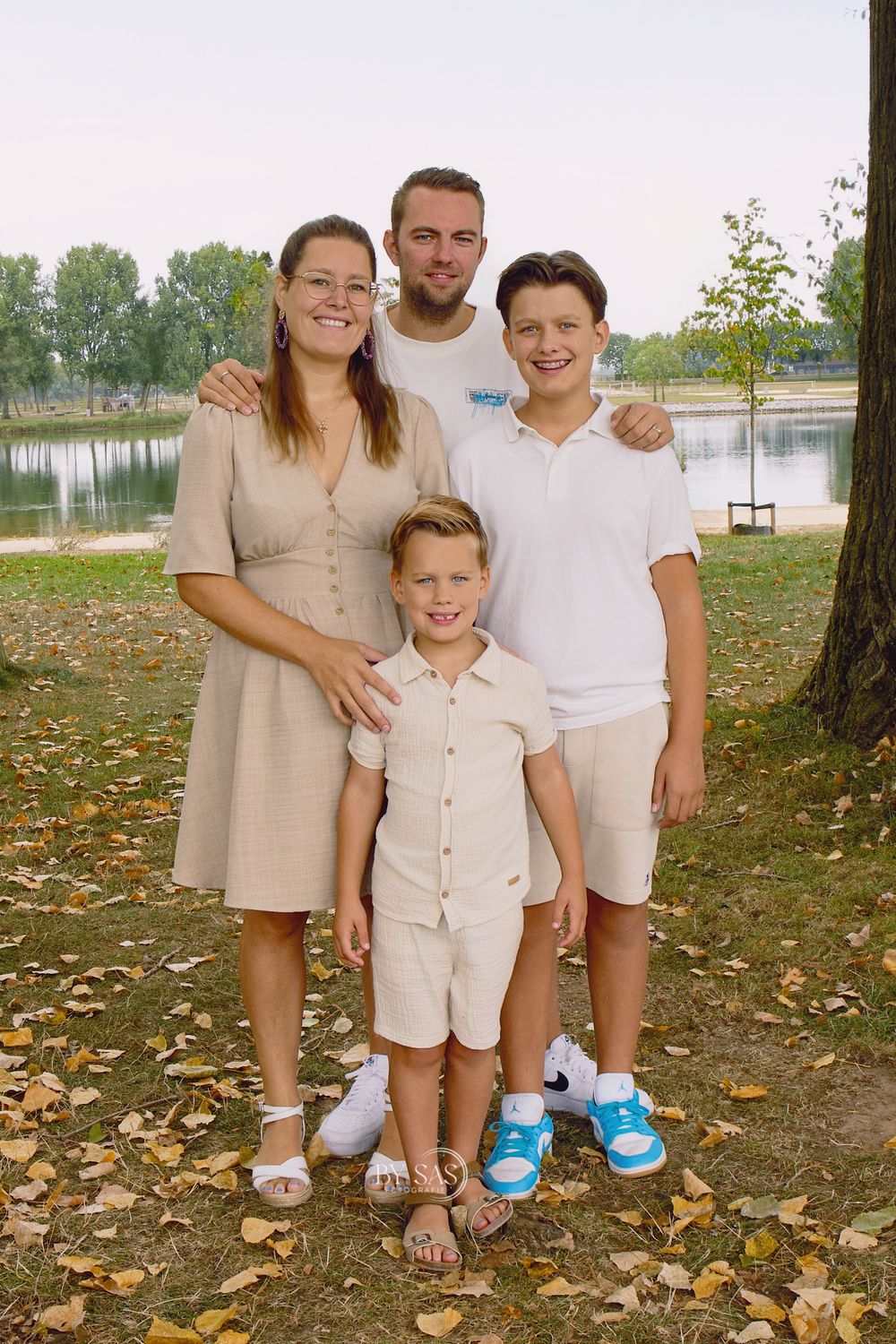 Family of four smiling together in a park near a lake with autumn leaves on the ground.