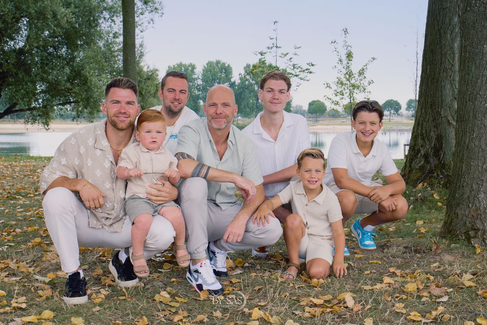 Family portrait of seven people posing outdoors near a tree and lake on a sunny autumn day with fallen leaves.