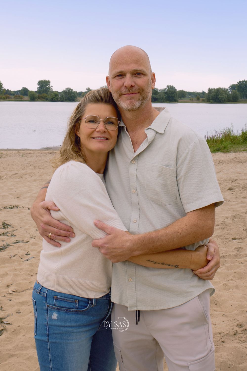 Smiling couple embracing on a sandy beach near a lake, woman in jeans and glasses, man in light shirt.