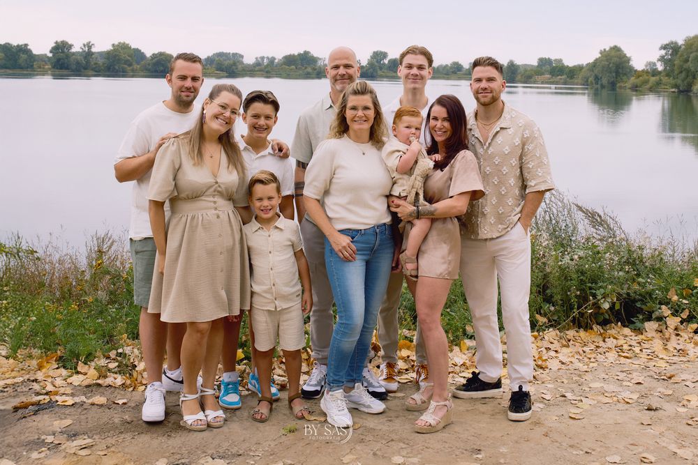 A large family group poses together smiling near a lake on an autumn day, dressed in coordinating neutral tones.