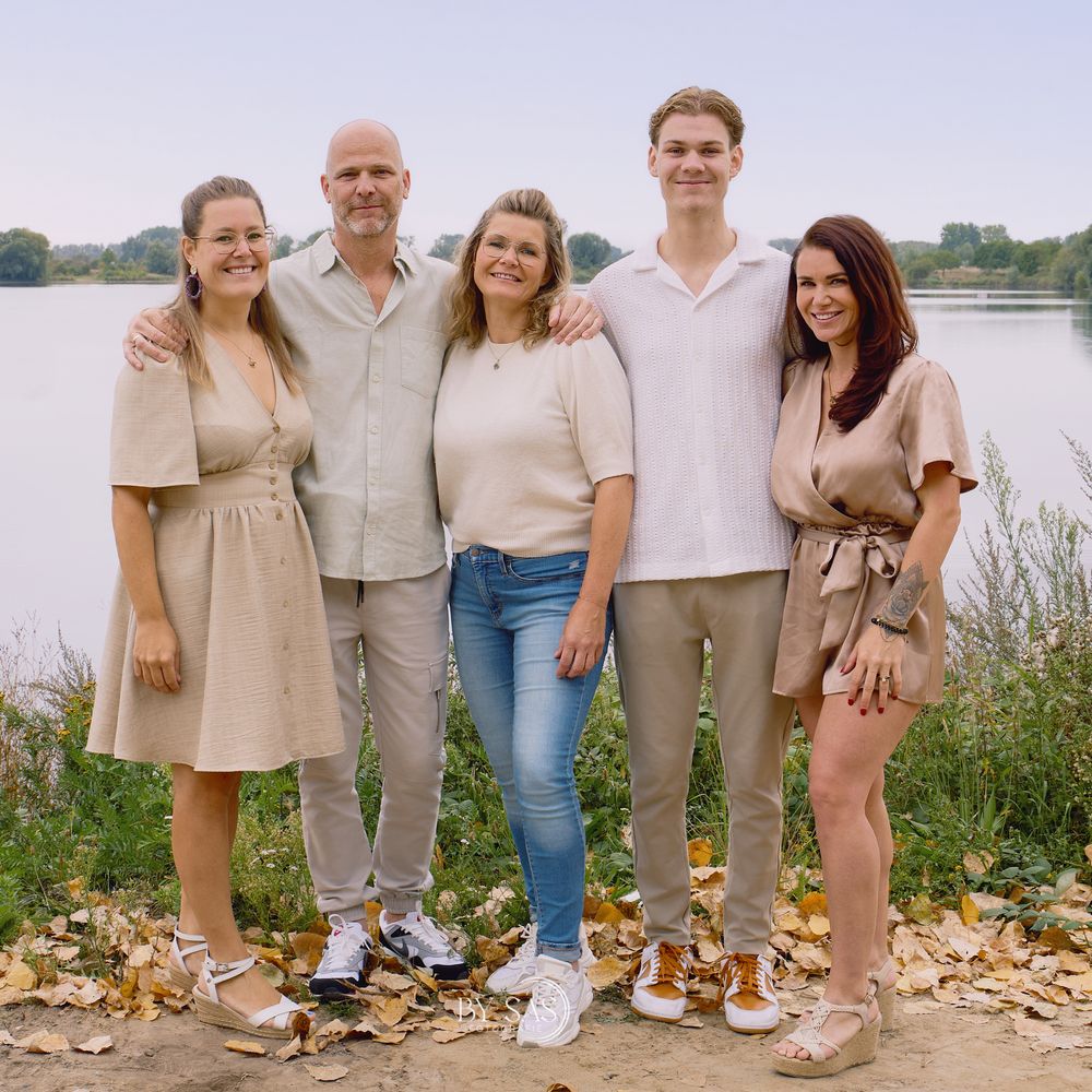 Family of five posing outdoors near a lake surrounded by autumn leaves in neutral toned outfits.