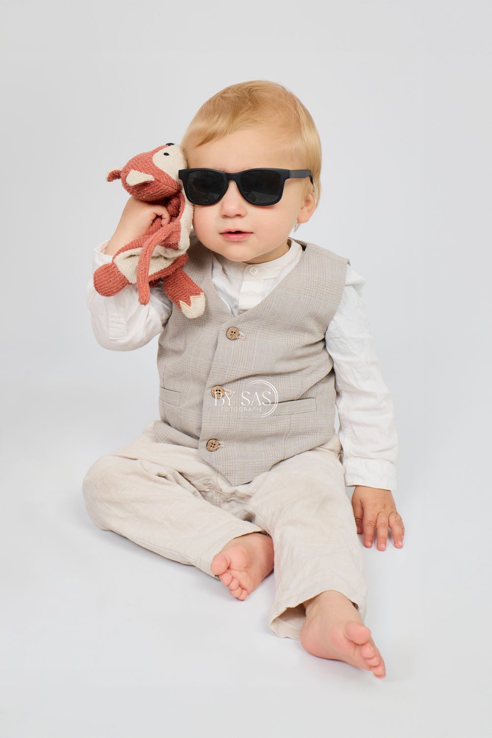 Stylish baby boy in sunglasses and beige vest suit holding a knitted fox toy, sitting on white background.