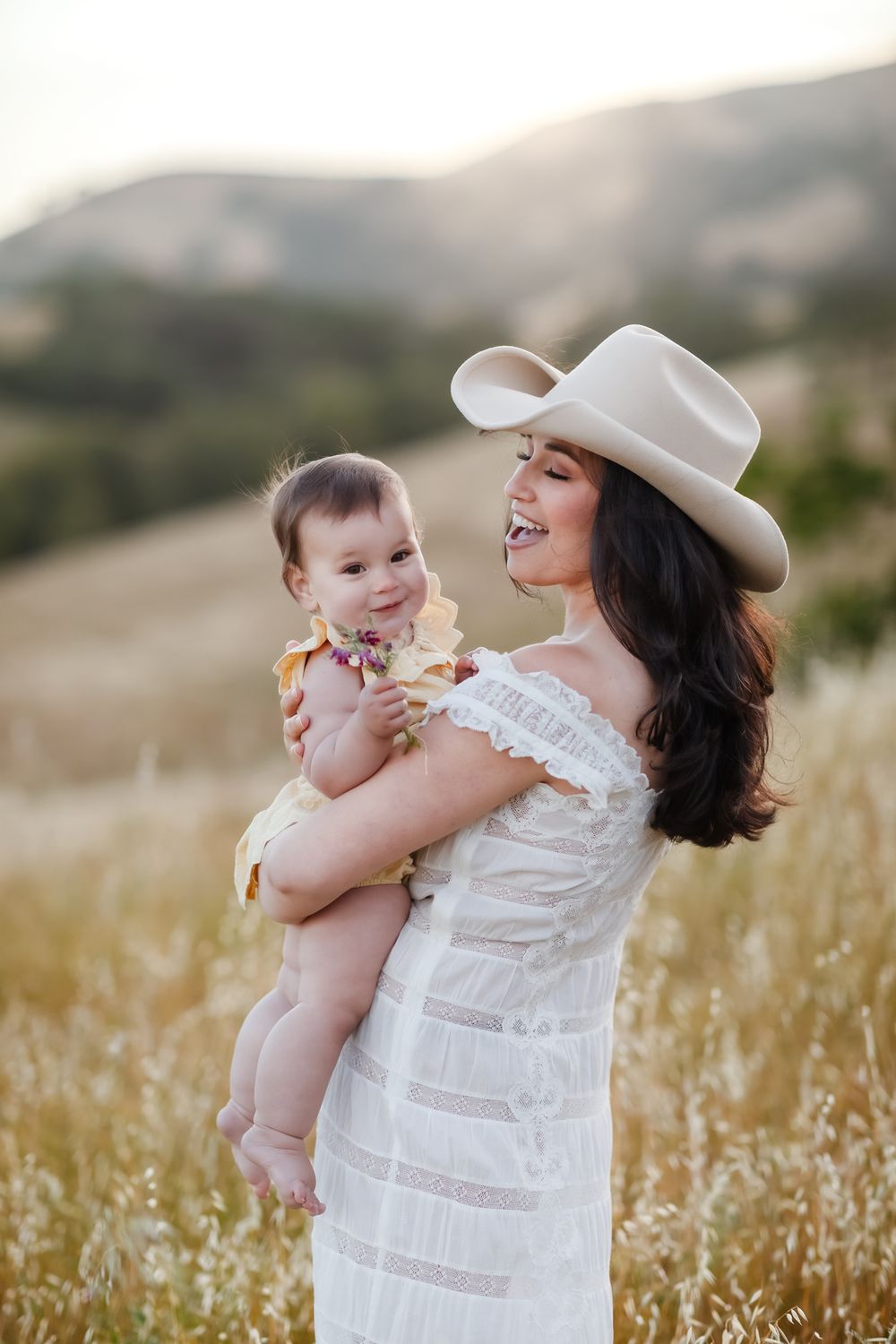 Happy baby girl in pale yellow is held by mother wearing white lace dress and cowboy hat in golden wheat field. Hampton family photographer