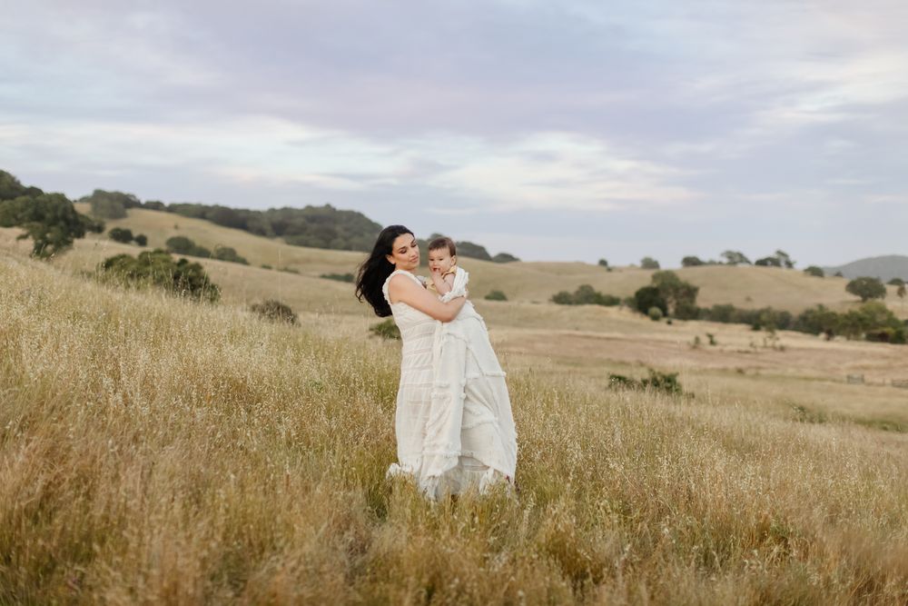 Mother in white lace dress holds baby walking through rolling golden field hills on cloudy day. Concord York family photographer