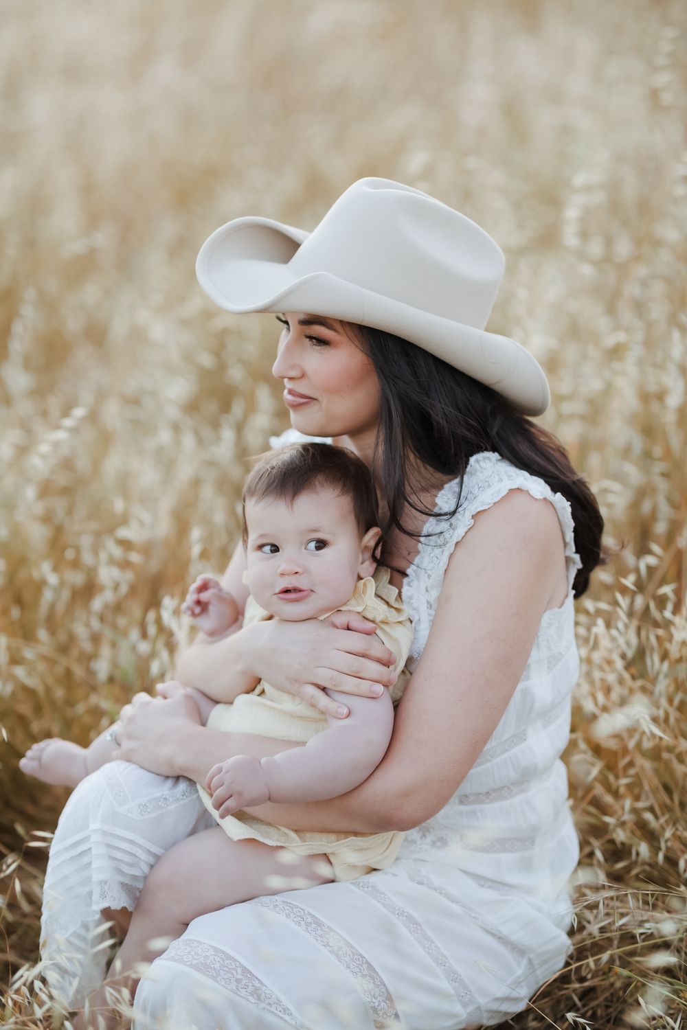 Dark hair mother in cream lace dress and cowboy hat holds baby wearing yellow in a golden field. North Shore motherhood family photographer