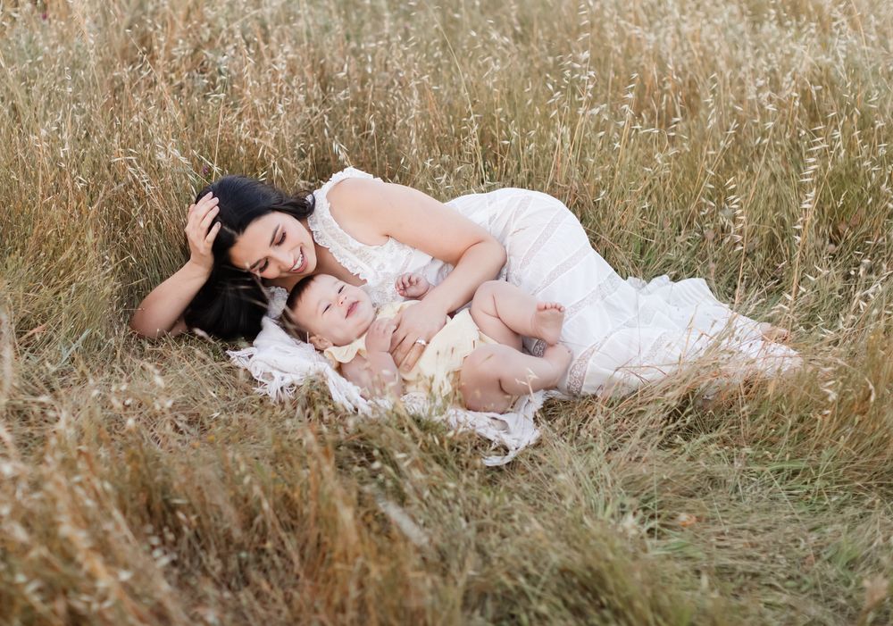 Dark-haired mother in white lace dress lays with laughing baby girl wearing yellow in golden grass. Wolfeboro Alton family photographer