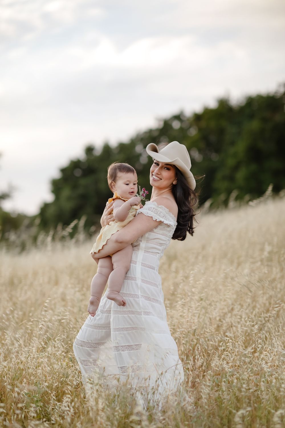Mother in white lace dress and cowboy hat holds baby girl in yellow standing in golden grassy field. Chelmsford Rye family photographer