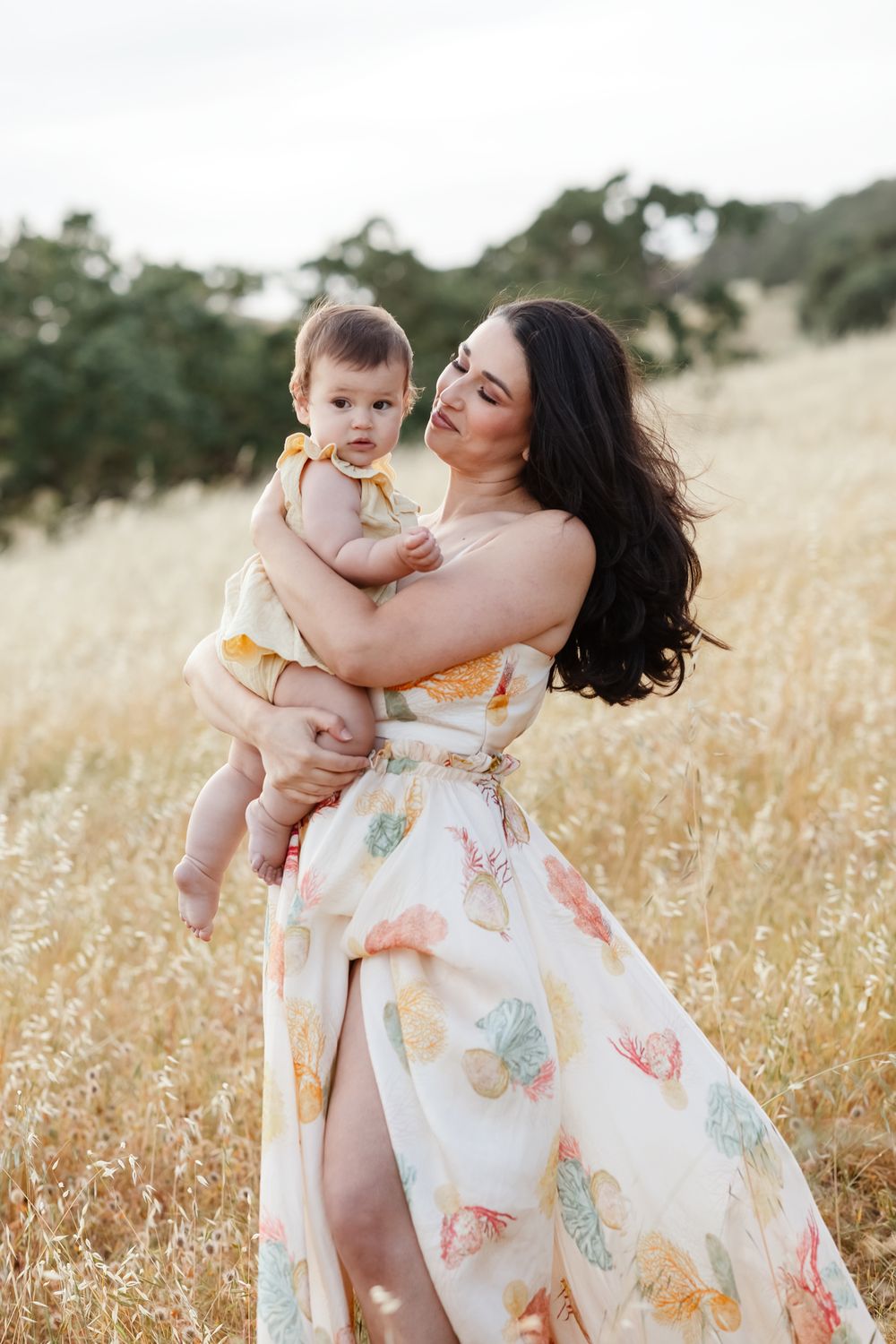 Mother in white seashell patterned gown holds baby girl in golden field for motherhood photos. White Mountains family photographer.