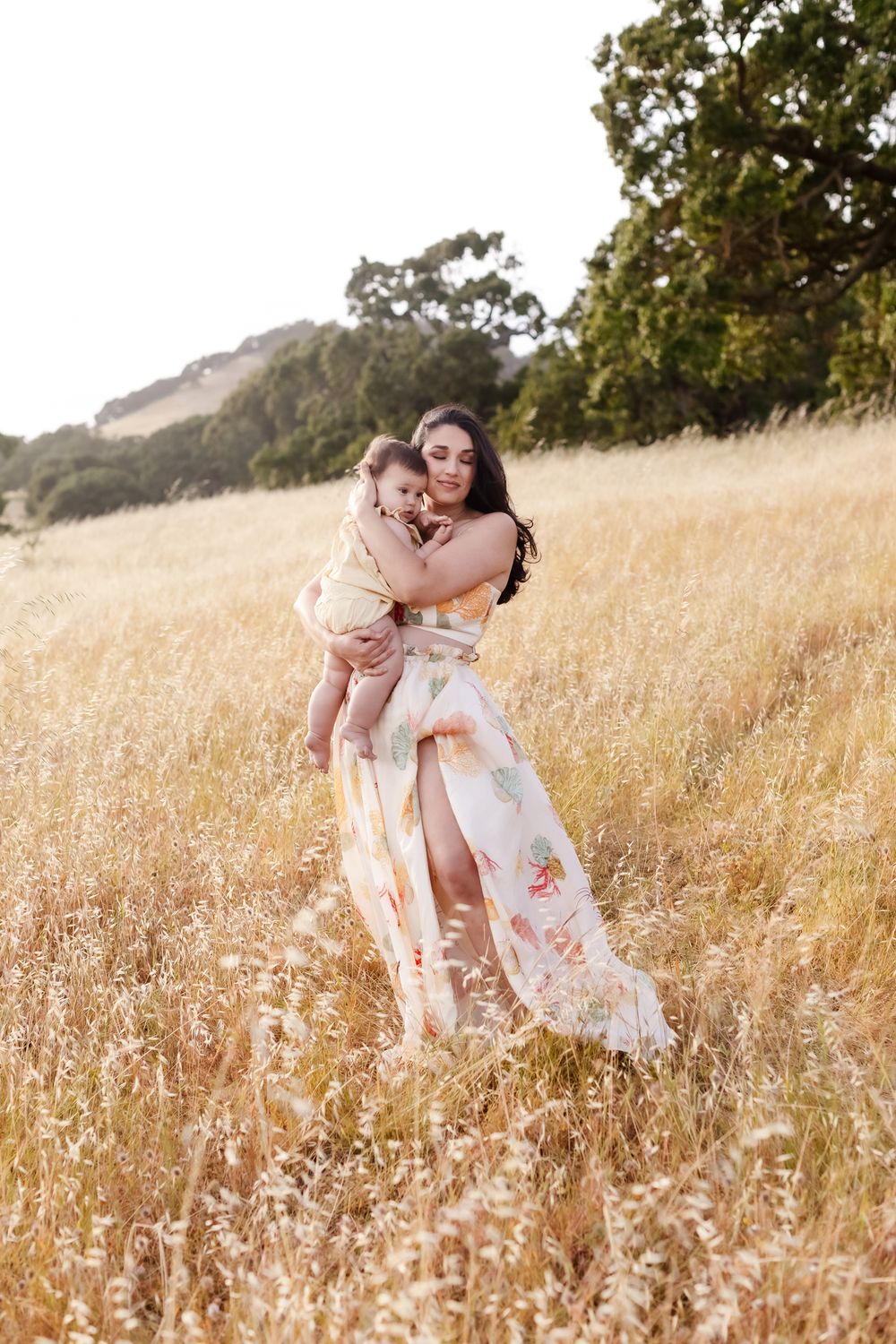 Mother in seashell patterned white dress embraces baby girl in golden wheat field. Concord Hampton Gloucester family photographer