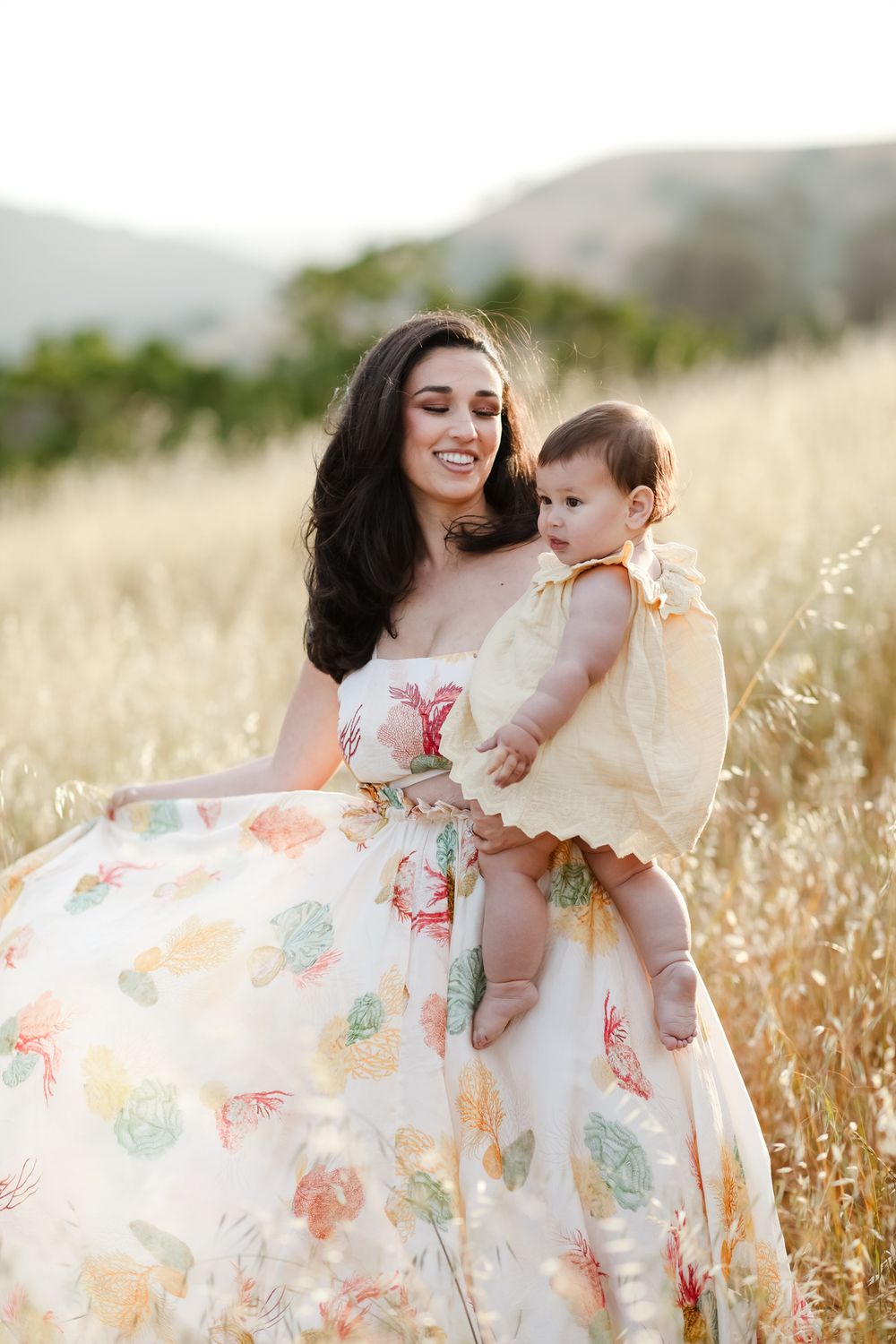 Smiling dark hair mother sways her dress as she holds baby girl wearing pale yellow in golden wheat field. Southern Maine family photographe