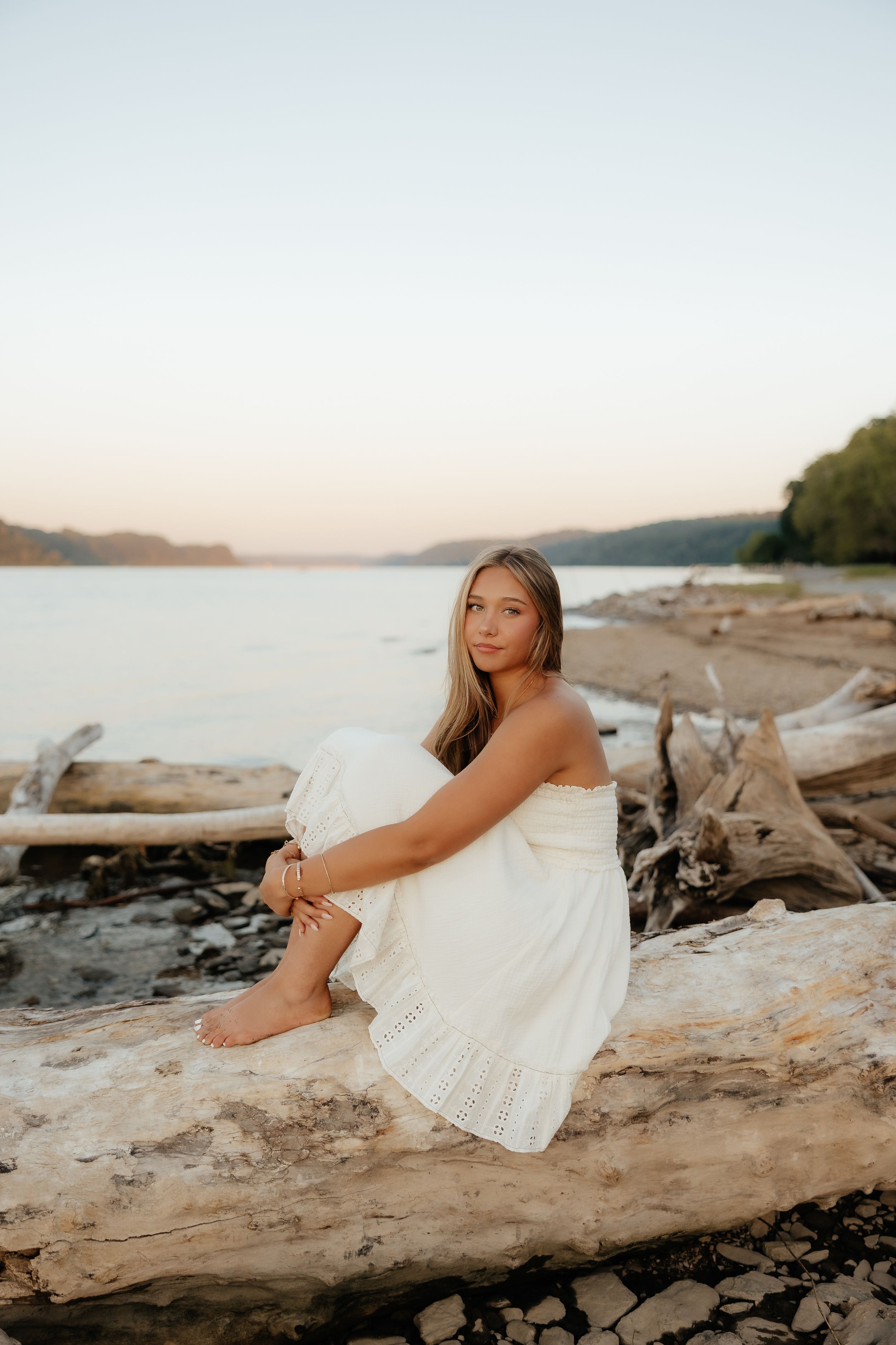Young woman in white dress sitting on driftwood by a serene lake at sunset, gazing peacefully into the distance.