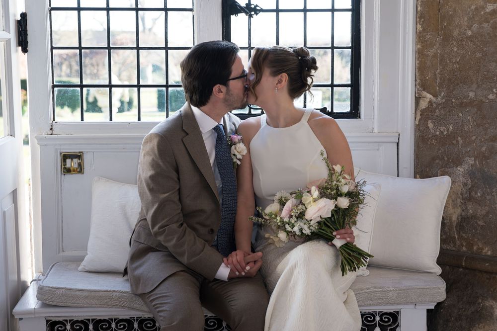 Bride and groom share a kiss while seated on a white bench, bride holding a pink floral bouquet near elegant windows.