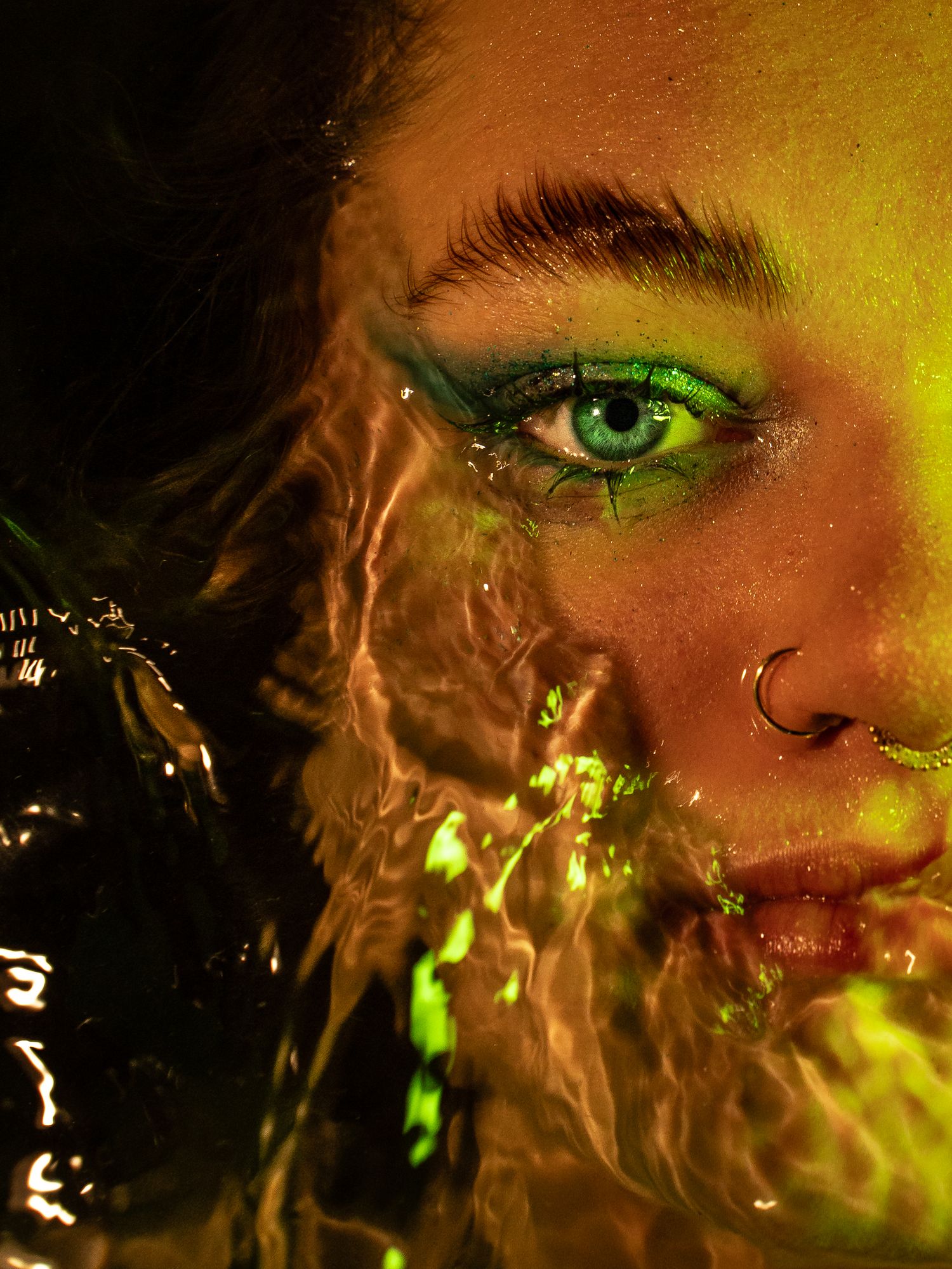 Close-up portrait of a woman with vibrant green eye makeup and nose ring, illuminated by neon green light.