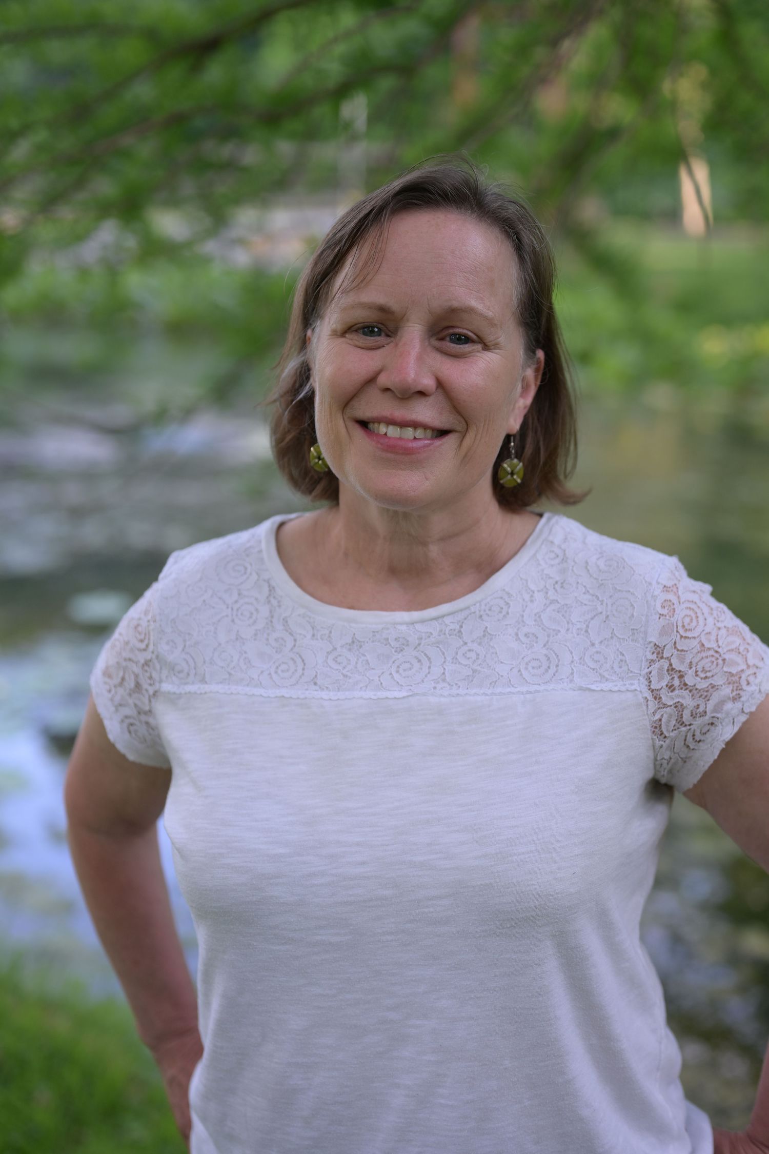 Smiling older woman with white curly hair wearing a white button-up shirt outdoors in natural green setting.