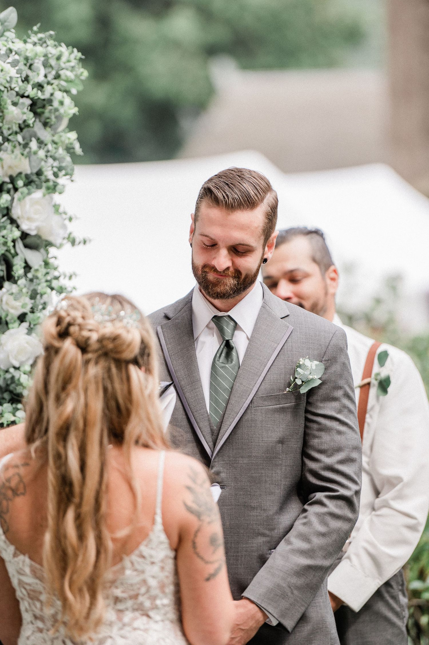 Groom in gray suit with green tie smiling emotionally during outdoor wedding ceremony beside floral arch.