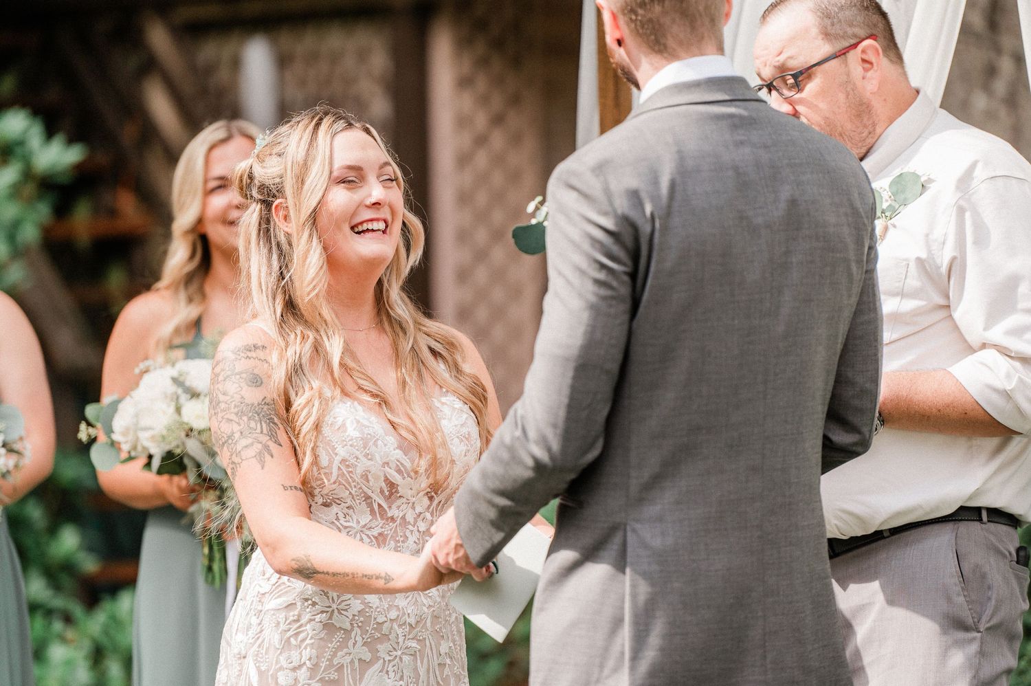 Bride smiling joyfully at groom during outdoor wedding ceremony, holding white bouquet, with bridesmaids in sage green dresses.