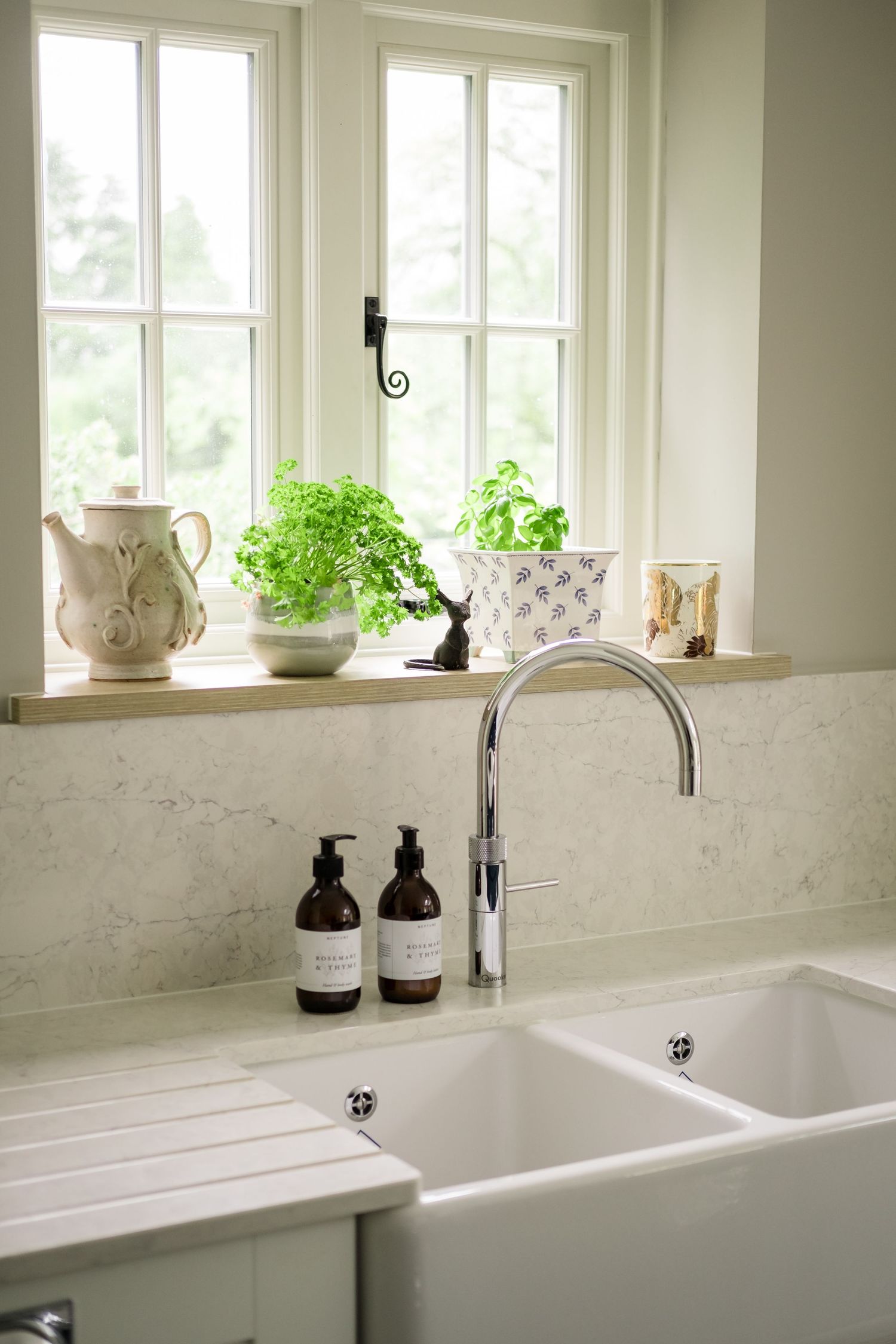 White farmhouse kitchen sink with chrome faucet, marble backsplash, and herb plants on windowsill in bright natural light.