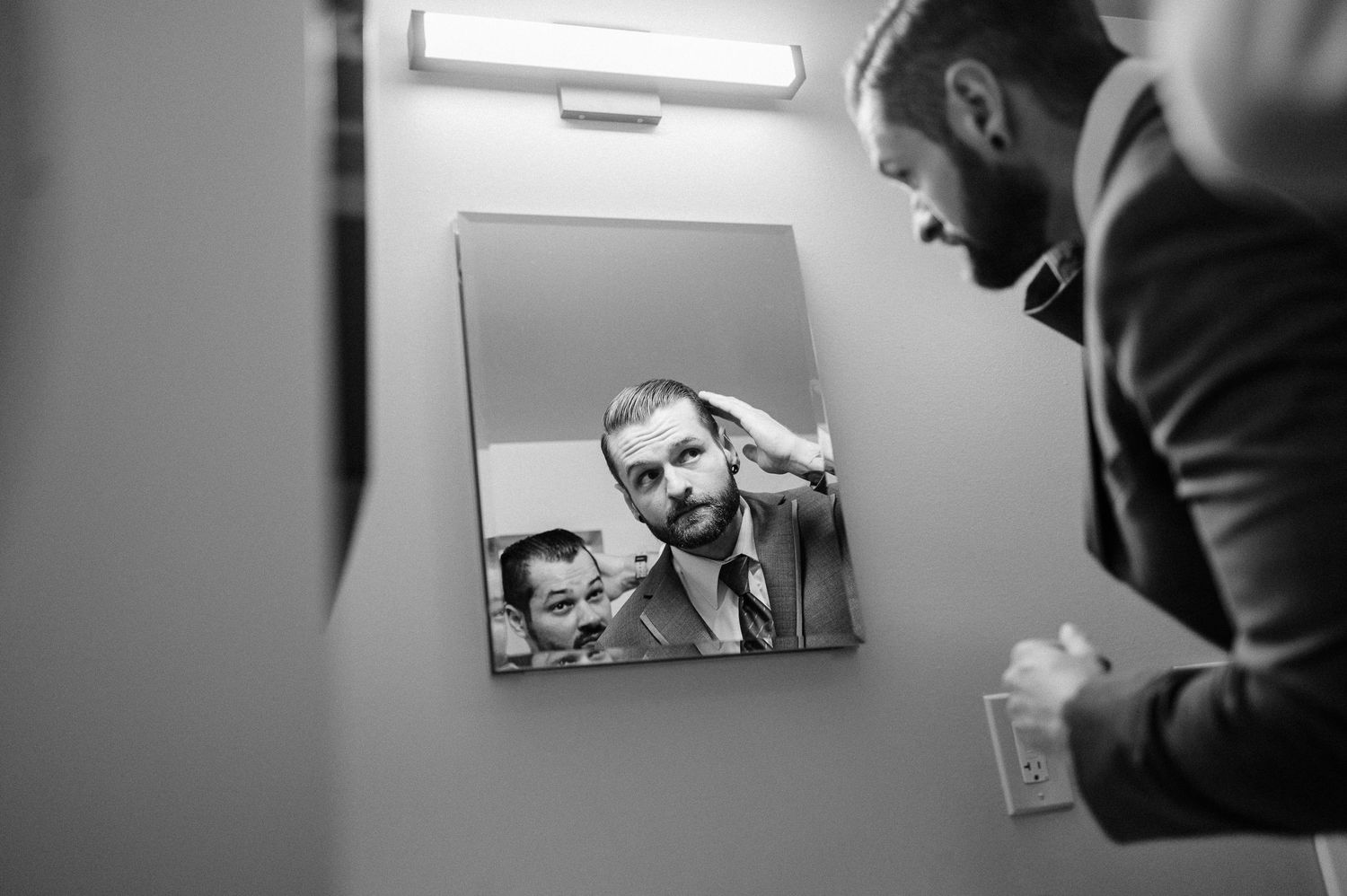 Black and white photo of a groom adjusting his hair in a bathroom mirror before his wedding ceremony.
