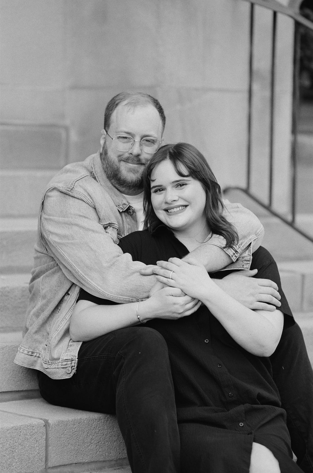 A couple sits on stone steps embracing warmly, smiling in a black and white engagement photo.