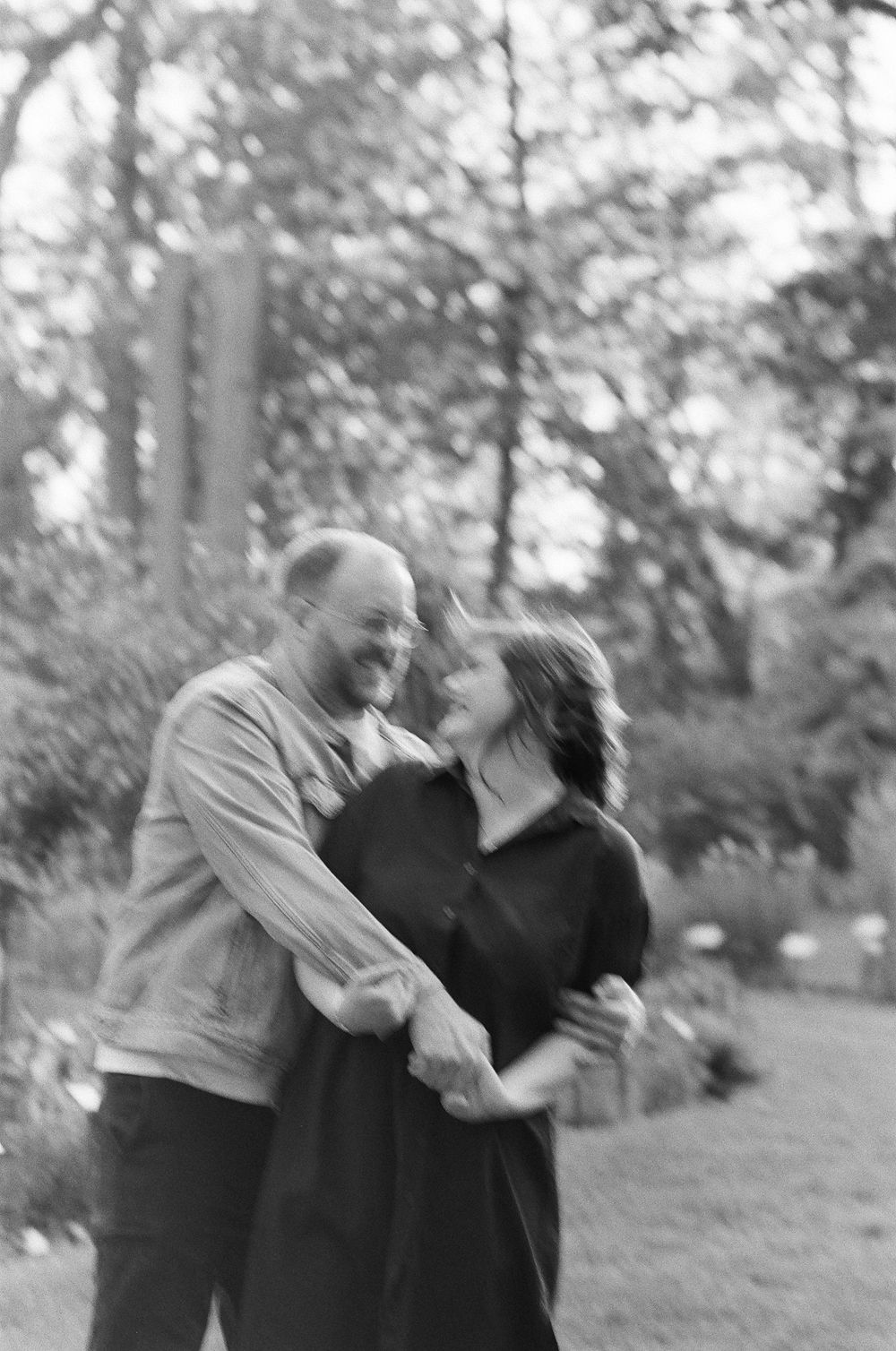 Black and white photo of a couple laughing and dancing together outdoors in a lush garden setting.