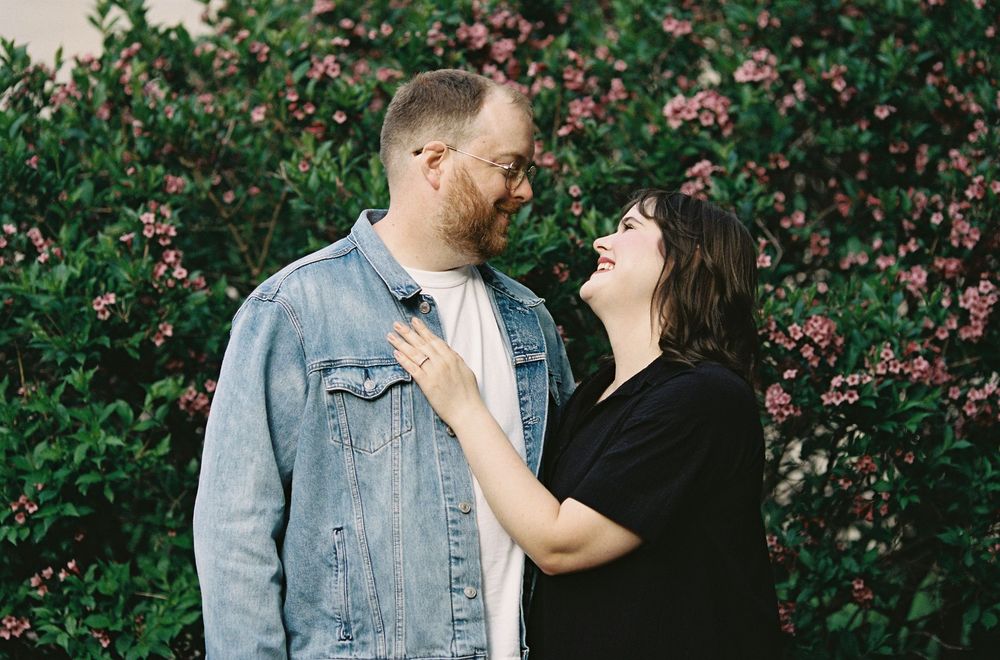 Couple sharing a tender moment in front of blooming pink flowers, woman in black dress touches mans denim jacket.