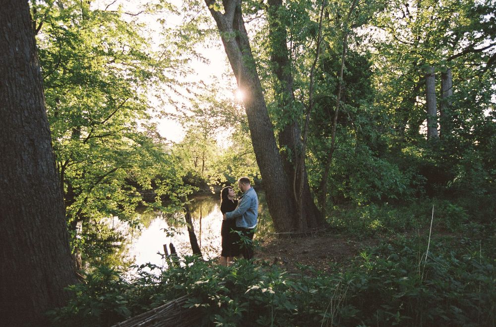 Couple embracing near a sunlit forest pond surrounded by lush green trees.