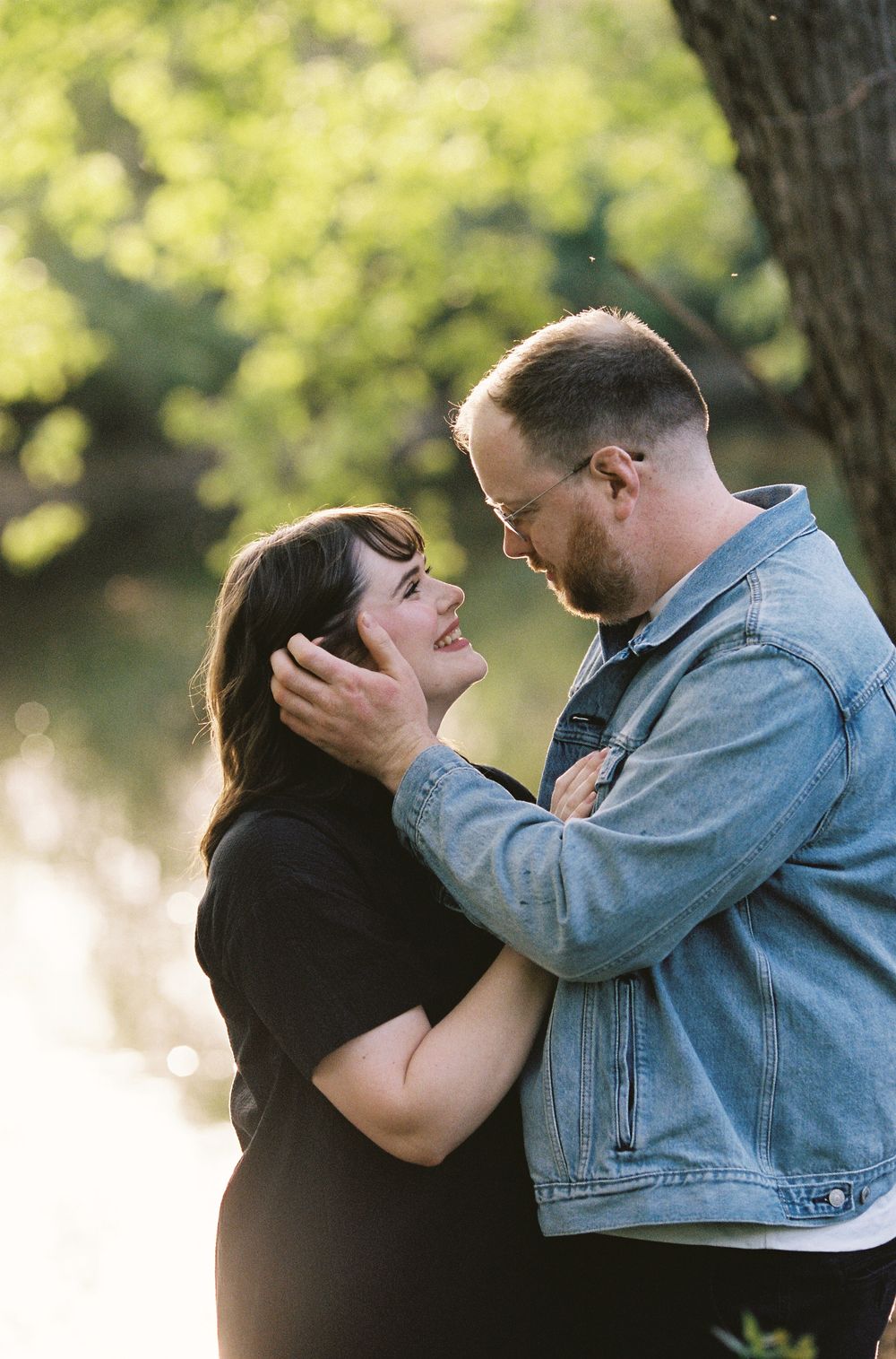 Couple sharing a tender moment outdoors, man holding woman's face near a sunlit pond.