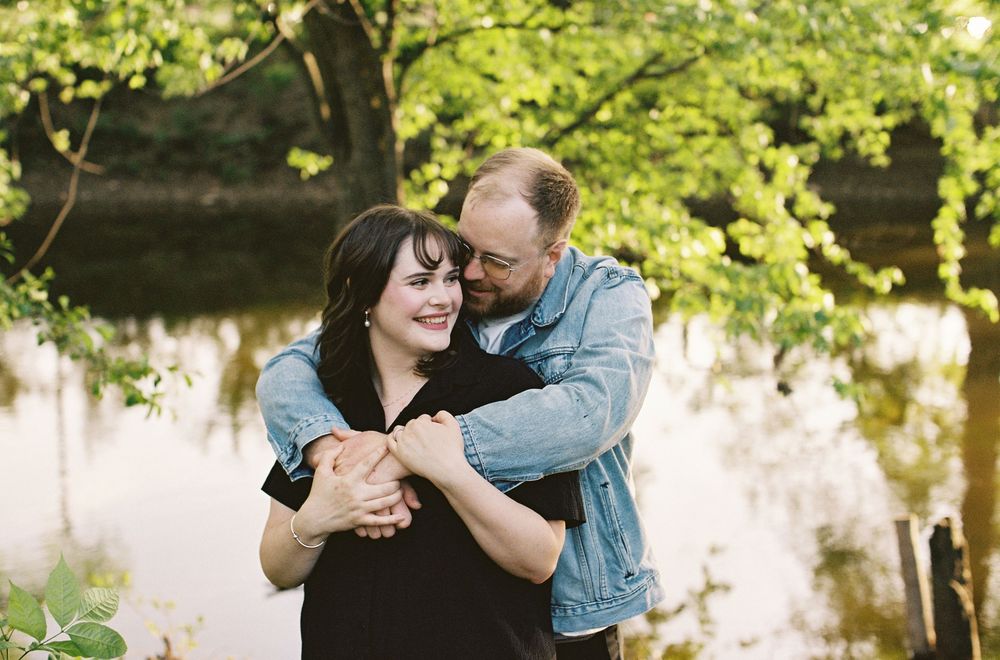 A couple embracing and smiling near a serene pond surrounded by vibrant green spring foliage.