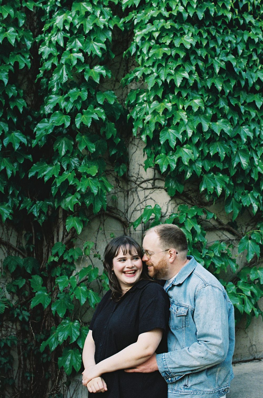 Smiling couple embracing in front of a wall covered in lush green ivy.