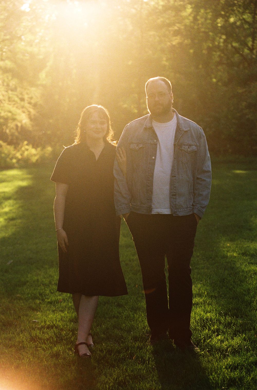 Couple standing together on green grass in golden hour sunlight with trees in background.