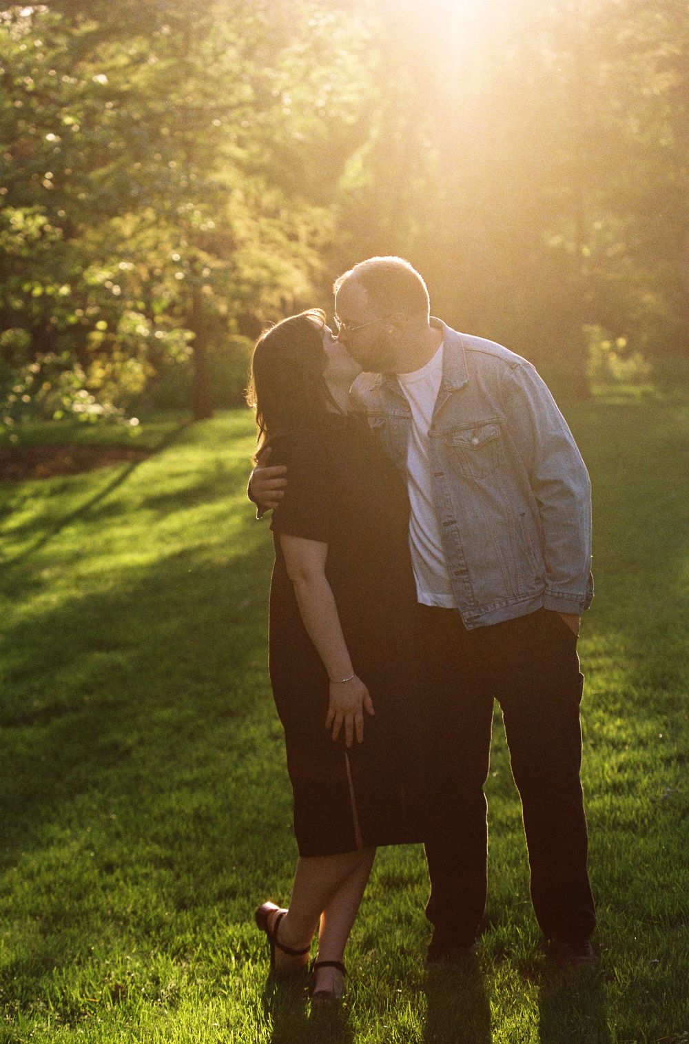 A couple sharing a kiss in a sunlit park with golden hour light glowing behind them.