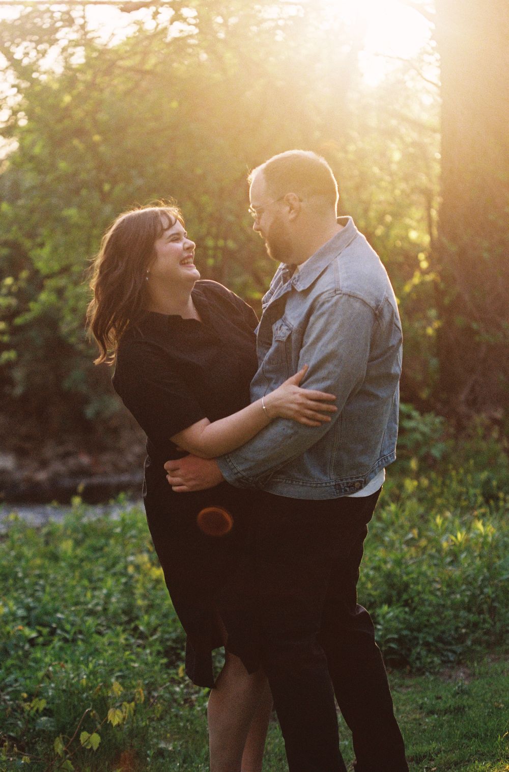 Couple laughing and embracing in golden sunset light surrounded by lush green trees.