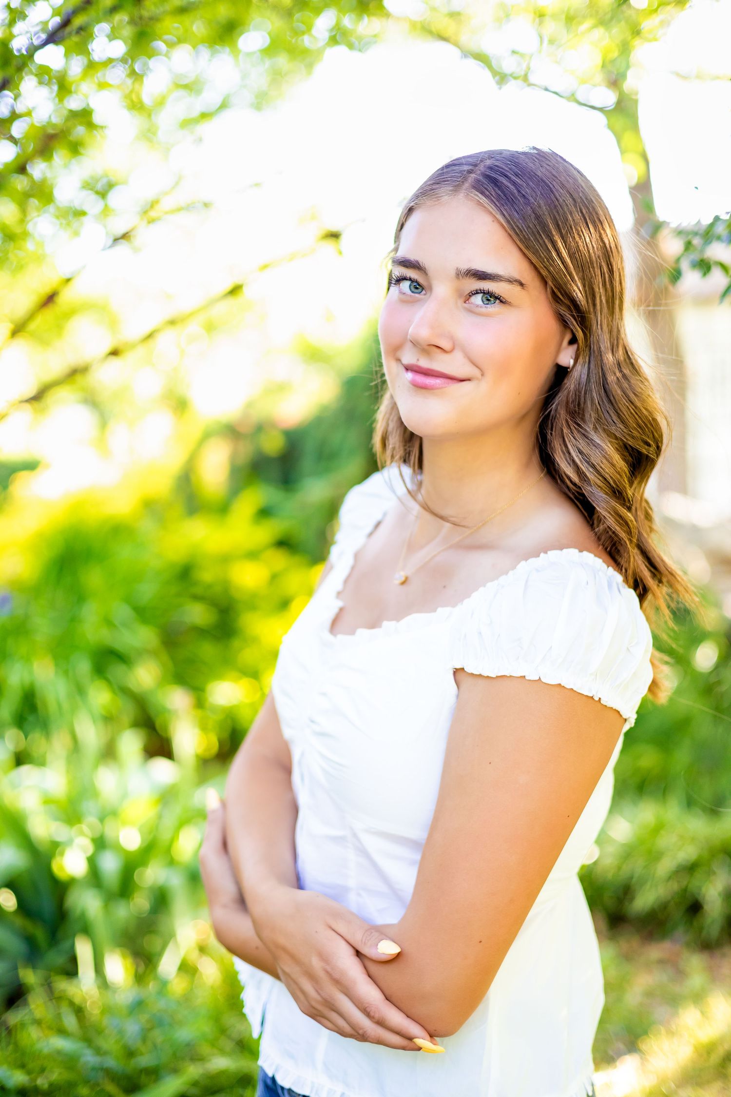 Young woman in white top smiling outdoors with lush green trees in sunlit background.