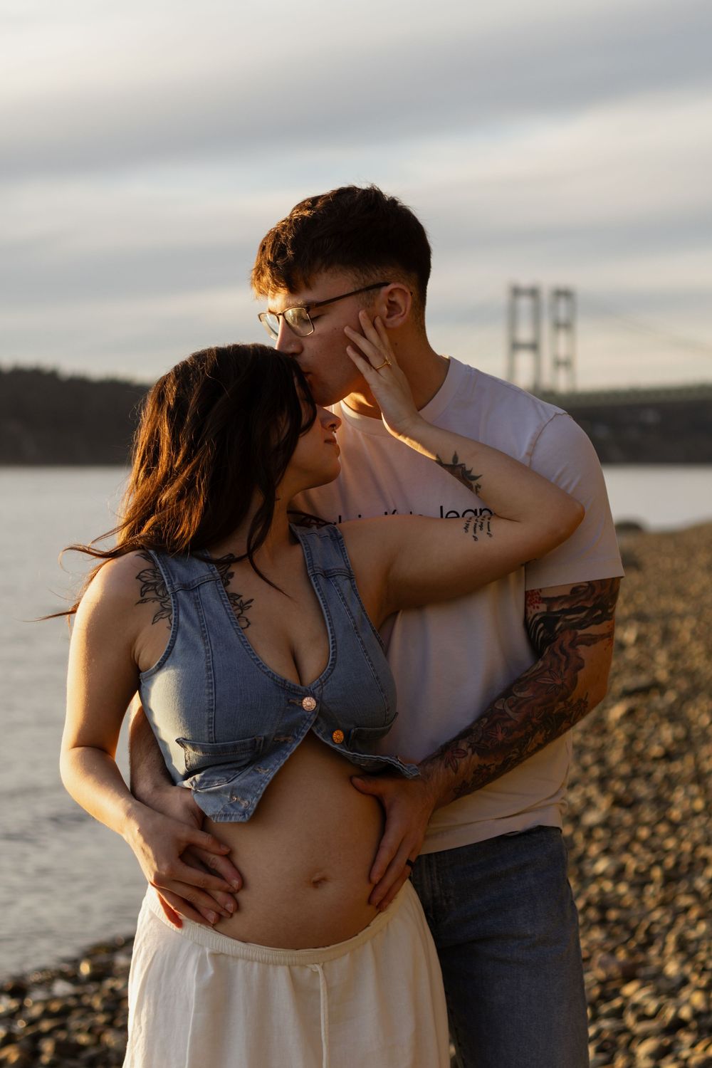 Tattooed couple sharing a romantic kiss on a rocky beach at sunset, with hood canal bridge and calm water in the background.