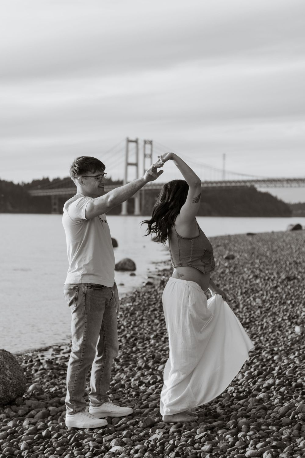 Black and white photo of a couple dancing on a rocky beach with hood canal bridge in the background.
