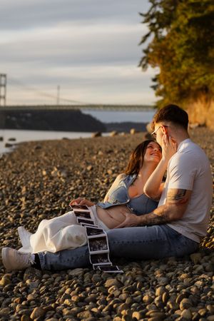 Couple sitting on rocky beach at sunset holding ultrasound photos, celebrating pregnancy announcement with hood canal bridge in background