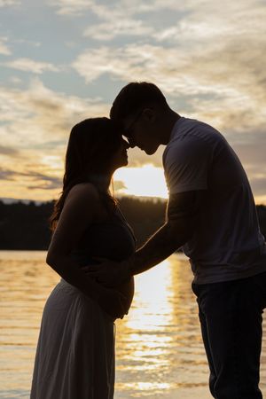 Silhouette of a pregnant couple touching foreheads at sunset by a lake, golden light reflecting on water.