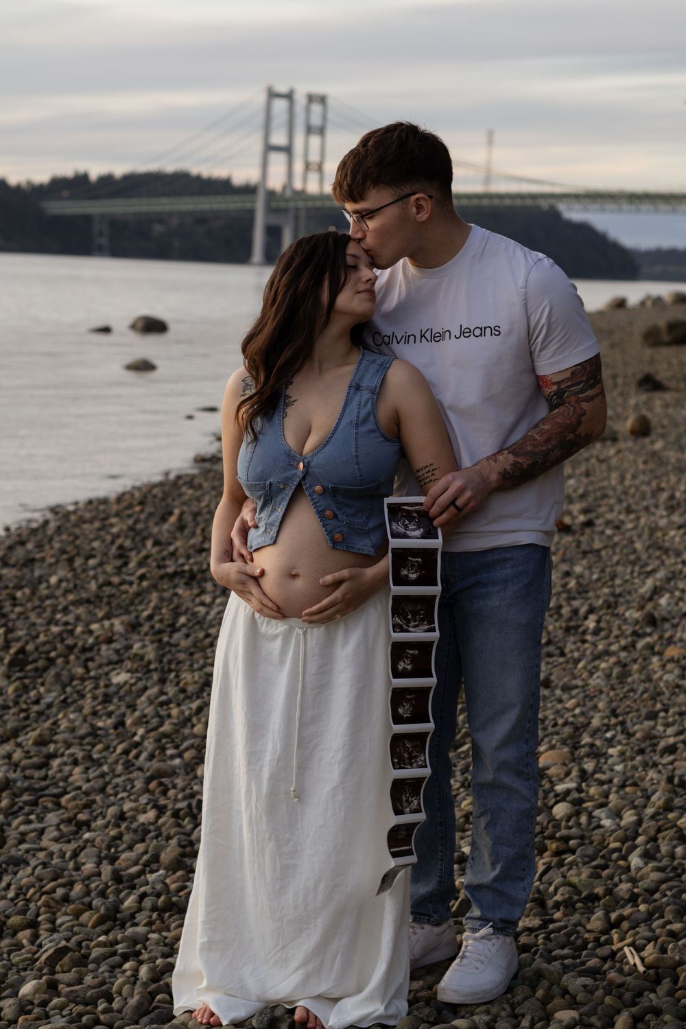 Pregnant couple embracing on rocky shore, holding ultrasound images with hood canal bridge in background.