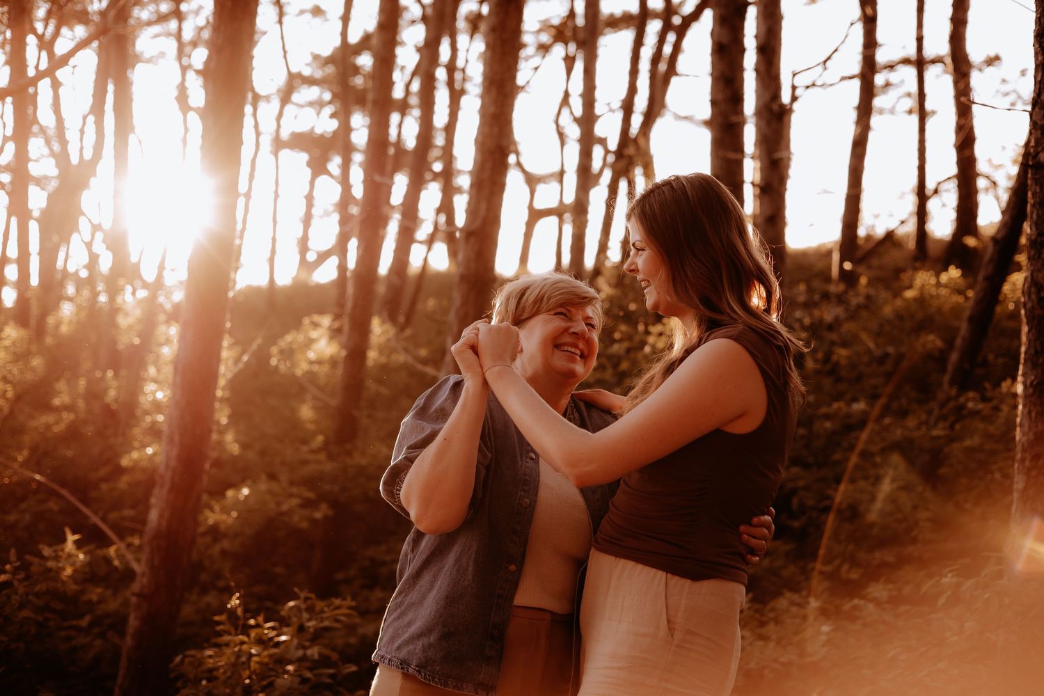 Two women embrace and smile warmly at each other in a sunlit forest during golden hour.