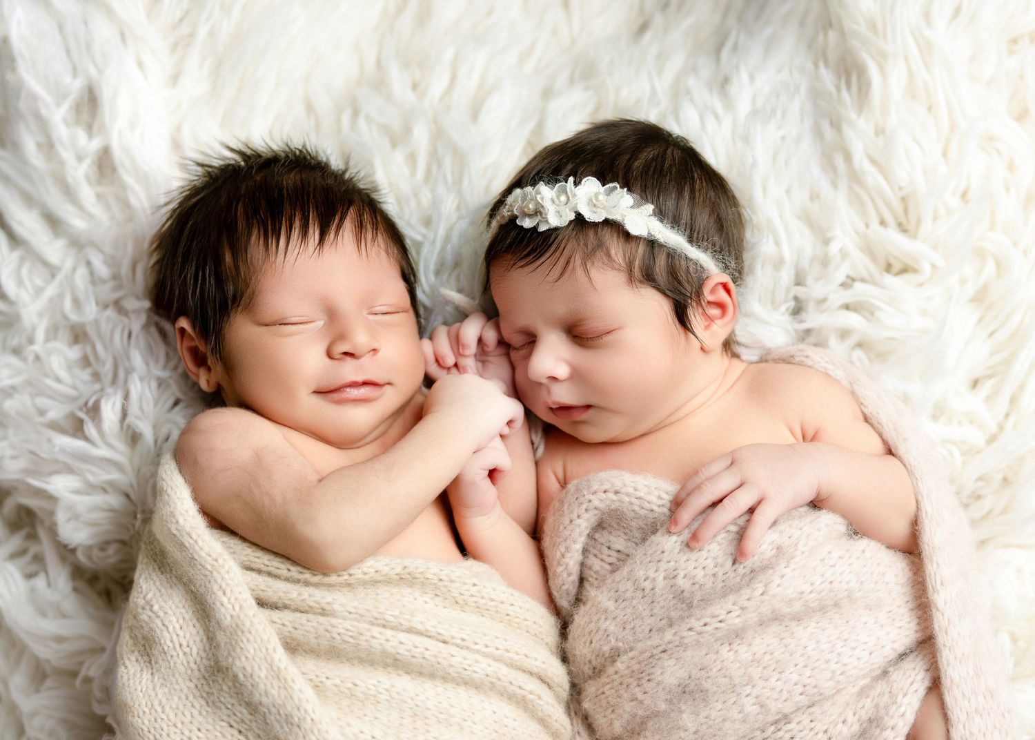 Two sleeping newborn twins wrapped in soft blankets on a white fur rug, one wearing a floral headband.