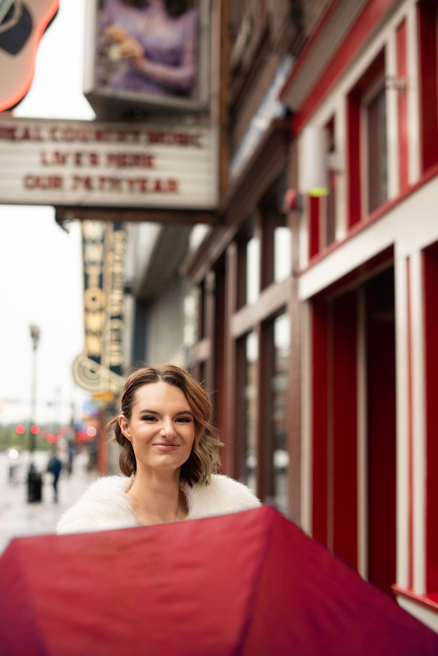 Woman smiling on Nashville Broadway sidewalk near country music venue marquee, holding red fabric, wearing white fur wrap.