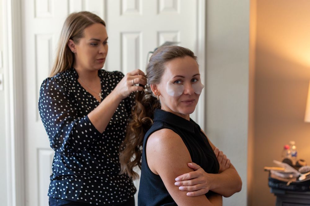 A woman styling another woman's curly hair indoors, both dressed elegantly for a special occasion.