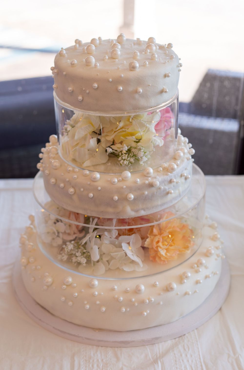 Elegant three-tier white wedding cake adorned with pearl details and fresh flowers between clear acrylic separators.