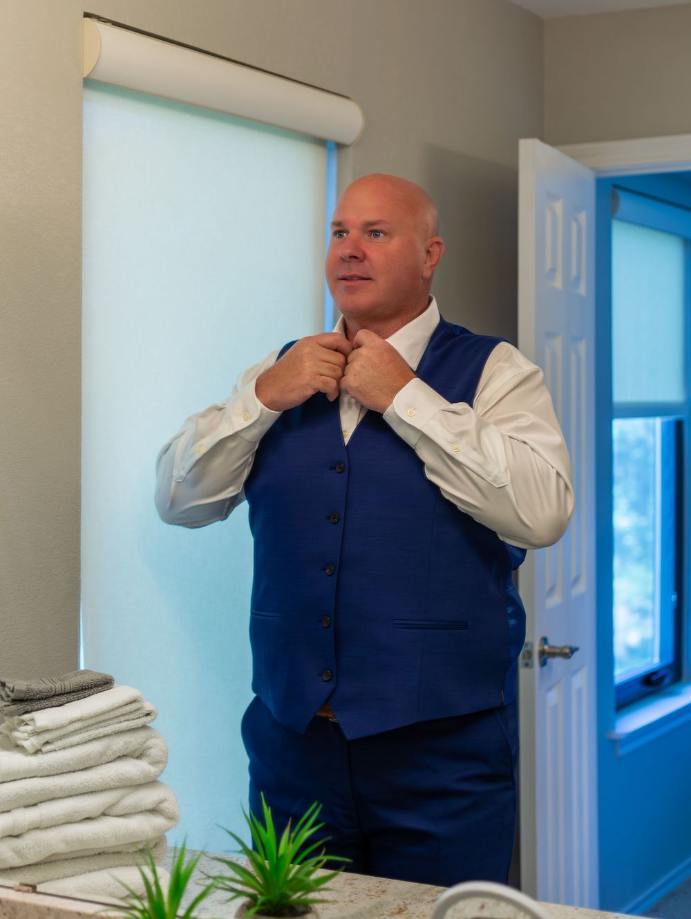 Bald man in a blue vest adjusting his tie while getting dressed in a bright room.
